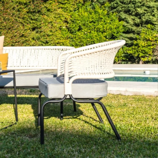 sillón de caño negro y cuerda beige con almohadón gris sobre el pasto junto a una piscina. Hay una mesita lateral negra que sostiene una bandeja con refrescos y se ve un sillón de dos cuerpos a s lado