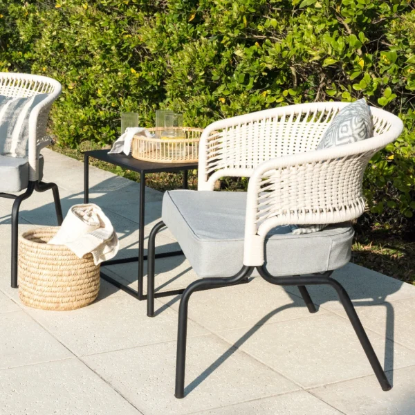 sillón de caño negro y cuerda natural en respaldo con almohadón gris junto a una piscina. Hay una mesita lateral negra que sostiene una bandeja con refrescos y en el piso una cesta de ratán con revistas