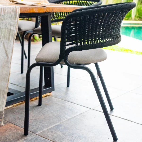 Silla de caño con almohadón en asiento gris y respaldo de cuerda trenzada negra. la toma es lateral y la silla se encuentra junto a una mesa